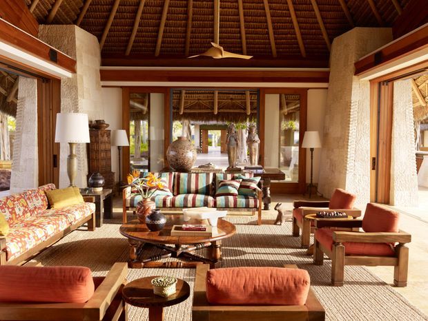 A spacious tropical living room with an open pavilion-like layout, exposed thatched roof and timber rafters, textured light stone or plaster columns, extensive timber-framed glazing, and colourful patterned upholstered seating arranged around low wooden and woven tables. The room is warm, airy, and richly layered with natural materials and earthy terracotta accents.