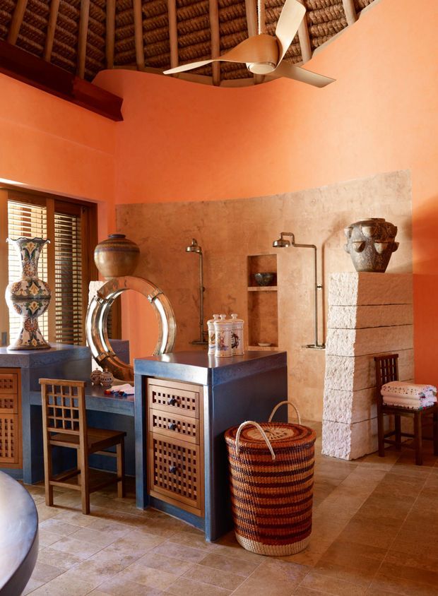 A richly styled rustic bathroom or dressing space with terracotta-orange plaster walls, a textured stone feature wall, exposed woven timber ceiling, and a ceiling fan. The room contains a dark timber vanity with lattice-front drawers, a round metal-framed mirror, wall-mounted basin fixtures, decorative pottery, woven storage, a tiled floor, and a carved stone fireplace-like feature. Warm natural light enters through timber shutters.