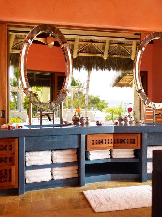 A tropical bathroom vanity with a long dark blue-grey stone counter, twin large circular framed mirrors, open shelving with neatly folded white towels, and warm timber cabinetry with decorative lattice fronts. Terracotta-orange walls and tiled flooring create a warm, resort-like interior. Open glazing reveals a thatched outdoor structure, palms, and bright coastal surroundings.
