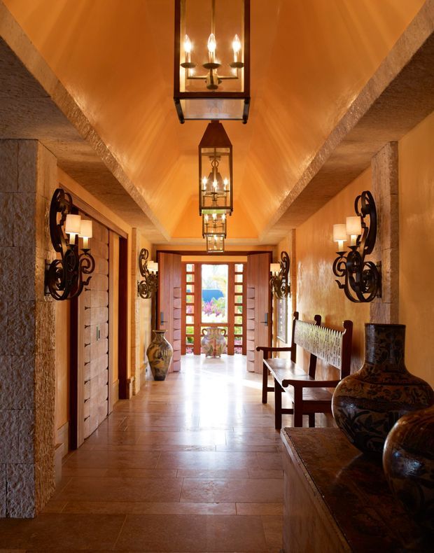 A long, warmly lit residential entry hall with terracotta-toned tiled flooring, textured stone columns and walls, exposed rustic ceiling beams, dark timber doors, and a series of suspended wrought-iron lanterns. Decorative wrought-iron wall sconces, carved timber seating, ceramic vessels, and a glazed timber entry door at the far end reinforce the rich, rustic character.