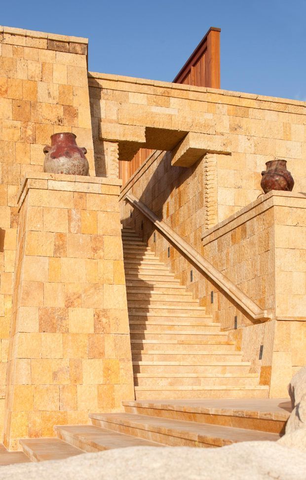 Sunlit exterior architectural view of a monumental stone staircase enclosed by warm golden-beige masonry walls. The stair has a timber handrail, projecting landings, decorative pottery urns, and a vertical timber architectural feature rising above the wall. The composition is strongly Mediterranean in material and form, set against a clear blue sky.