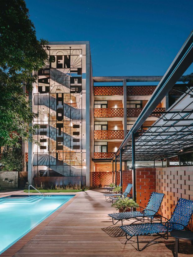 Night-time architectural view of a contemporary multi-storey building organized around a swimming pool and timber pool deck. The facade combines exposed concrete framing, red brick breeze-block screens, extensive glazing, and a prominent geometric mural or graphic facade panel. A lightweight steel-and-glass canopy covers the poolside seating area, with metal lounge chairs arranged beside the water.
