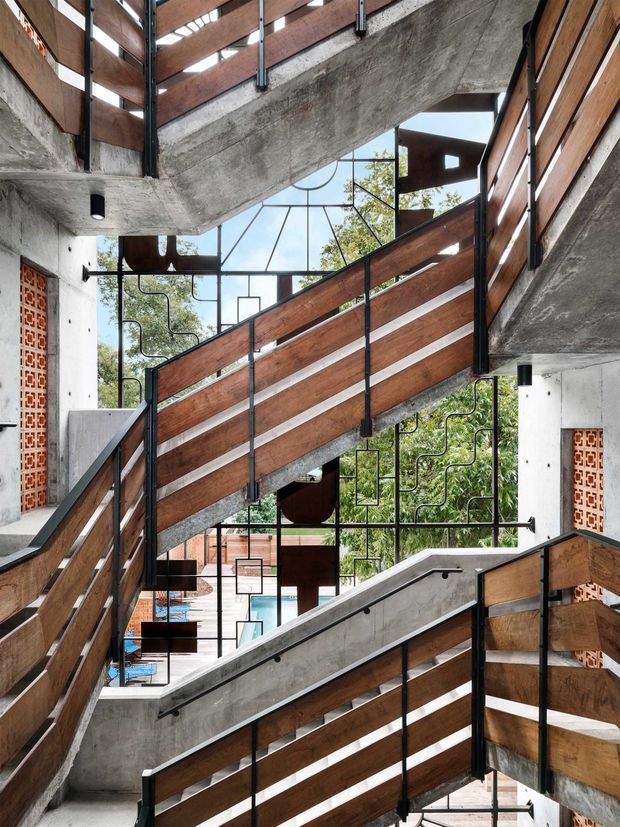A dramatic multi-level interior stairway with intersecting concrete stair flights, warm timber balustrade panels, black metal framing, and decorative geometric metal screens. Exposed concrete, red patterned brick screens, large glazed openings, and glimpses of greenery and a pool create an industrial yet warm architectural composition.