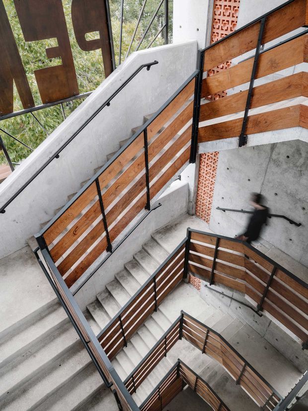 A multi-level interior stairwell with exposed concrete stairs and walls, black metal handrails and framing, and warm timber slat balustrades. A decorative terracotta breeze-block screen and large glazed openings add texture and natural light. The image is photographed from above, emphasizing the zigzagging stair flights and vertical circulation.