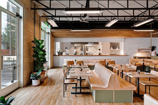 A spacious contemporary commercial dining space, likely a restaurant or café, with an open kitchen and service counter at the rear. The interior combines exposed warm brick walls, timber flooring and ceiling cladding, white subway-tile kitchen walls, black exposed steel ceiling structure, large steel-framed windows, linear pendant lighting, and built-in pale timber booth seating. Tables and chairs are arranged throughout the bright, airy room, with indoor plants adding greenery.