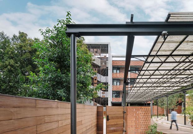 View from an outdoor covered entry or passage beside a contemporary building, featuring a dark steel pergola structure with translucent roofing, timber privacy fencing, exposed brickwork, and a paved walkway. Dense greenery and neighbouring multi-storey buildings are visible beyond.