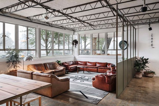 An expansive industrial-style living room or lounge with exposed steel roof trusses, polished concrete flooring, and full-height glazing. The space is furnished with several brown and rust-toned leather sofas and armchairs arranged around a large geometric rug and low coffee table. A large timber dining or work table occupies the foreground, while indoor plants, exposed services, and black-framed glazed partitions reinforce the warehouse-like character.