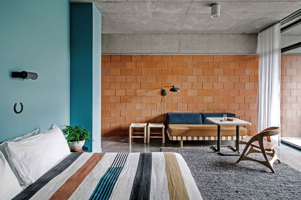 A stylish bedroom with a partially visible bed in the foreground and a compact sitting or dining area beyond. The room features exposed orange brick walls, a raw concrete ceiling, a bold teal painted wall and column, large corner glazing with sheer curtains, a blue upholstered banquette, a small table, timber chairs, and a textured grey rug. The aesthetic combines industrial architectural finishes with colourful mid-century-inspired furnishings.
