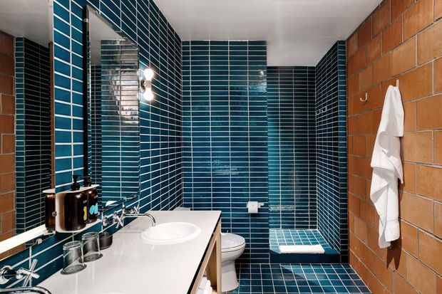 A compact bathroom with striking teal-blue rectangular tiled walls and floor, white grout, a white vanity with integrated basin, wall mirror, exposed chrome tapware, toilet, and a small tiled shower area. The right wall features warm orange-brown brick-style tiles with a towel hook and hanging white towels. The design has a bold, graphic mid-century-inspired character with strong colour contrast and reflective surfaces.
