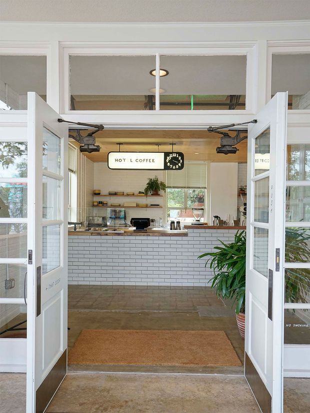 View through open glazed double doors into a compact coffee bar or café interior. The space features a white subway-tile counter front, timber countertop, open timber shelving, a hanging coffee sign and clock, warm concrete or stone flooring, and abundant daylight. White-trimmed doors and transom glazing create a traditional shopfront-like entrance, with plants framing the doorway.