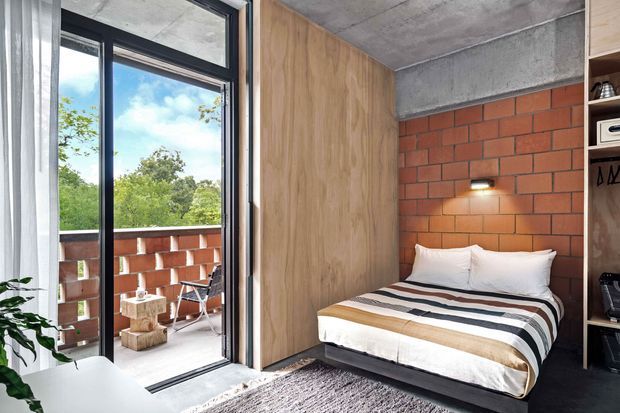 Compact industrial-style bedroom with a low platform bed, striped bedding, warm timber wall panelling, exposed concrete ceiling and beam, and a red brick feature wall. Full-height black-framed glazing opens to a small balcony, bringing in abundant natural light and views of greenery. Built-in dark shelving is visible at the right, while a textured rug and sheer curtains soften the space.