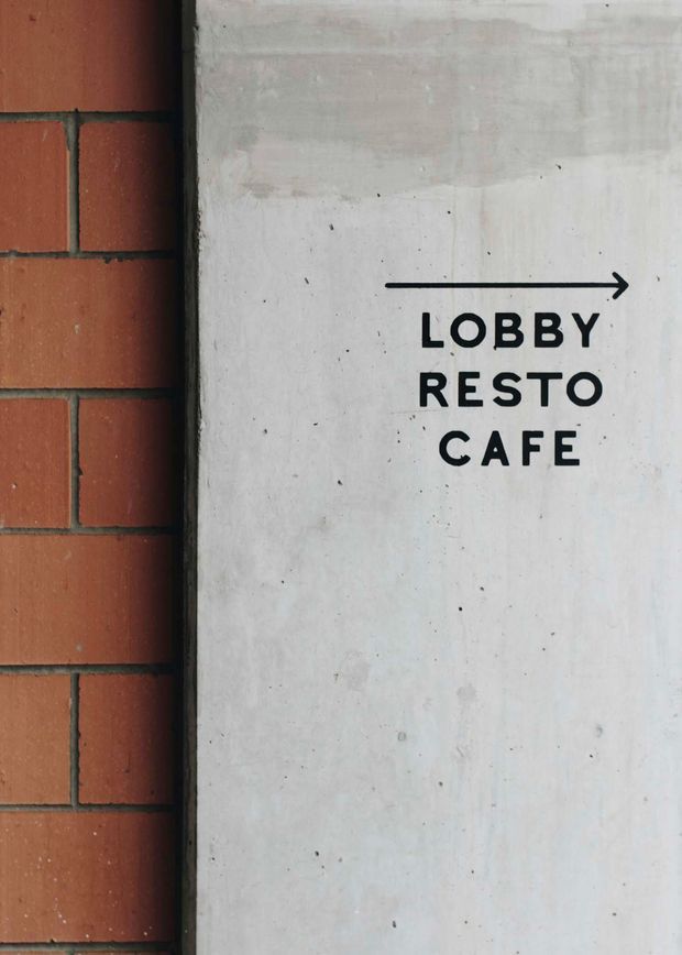 Close-up of a wayfinding sign mounted beside a red brick wall. The sign is a pale raw concrete surface with bold black lettering and a right-pointing arrow directing visitors to the lobby, restaurant, and cafe. The image has a utilitarian commercial and industrial character.