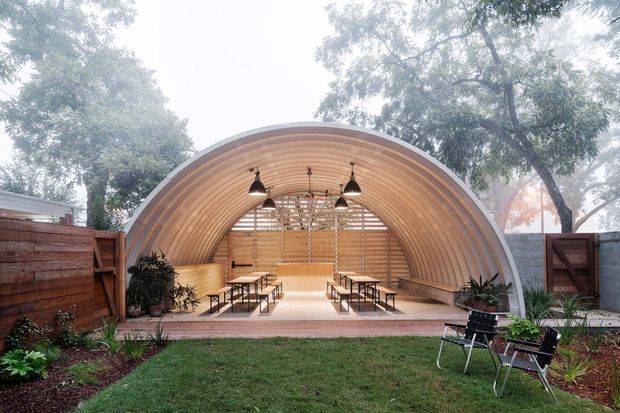 An outdoor communal pavilion with a distinctive curved, barrel-vaulted timber shell. The open-sided structure contains long picnic-style tables and benches, hanging pendant lights, and a rear timber screen. It is set within a landscaped garden courtyard with planting, lawn, fencing, and outdoor lounge chairs. The design has a warm, inviting, semi-enclosed gathering-space character.