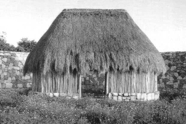 Black-and-white photograph of a small traditional thatched hut or shelter with a broad, steeply pitched organic roof, vertical timber or reed walling, and a low rough-stone perimeter wall behind it. The structure is set in an open grassy landscape.