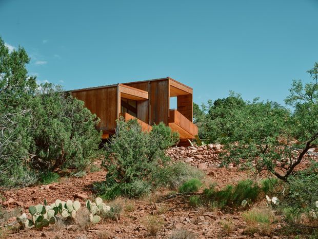 Exterior view of a compact modern timber-clad house set among rugged, rocky desert terrain and native scrub. The building has a boxy form with deep recessed openings, projecting balcony-like platforms, and a flat roof, surrounded by red rock, prickly pear, and drought-tolerant vegetation under a clear blue sky.