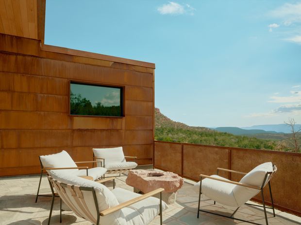 A sunlit rooftop terrace with rust-toned metal-clad walls and balustrades, furnished with four mid-century style lounge chairs and a rough stone coffee table. The terrace overlooks a dry, hilly landscape and distant mountains beneath a clear blue sky.