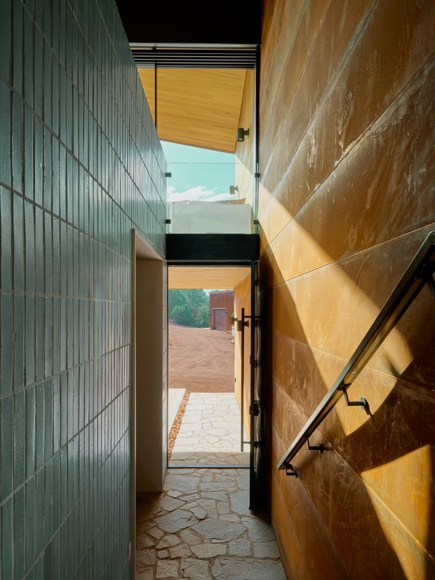 A narrow architectural passage or stair hall framed by contrasting tiled walls, with an irregular natural-stone floor, black metal handrail, timber soffits, and overhead glazing that brings in strong daylight. The passage opens toward an outdoor courtyard or driveway, creating a dramatic indoor-outdoor connection. The architecture has a contemporary, material-focused character with warm timber and copper-brown wall surfaces balanced by a dark blue-green tiled wall.