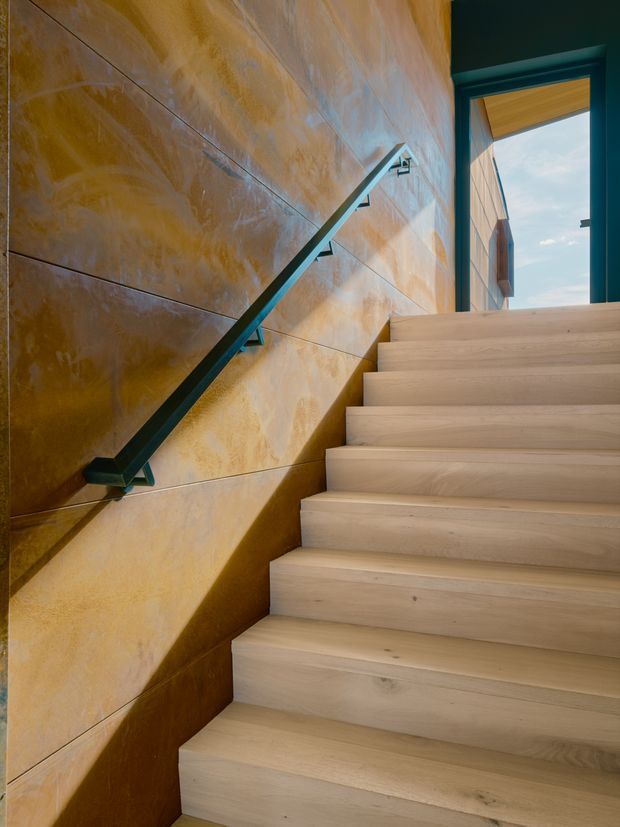 Interior view looking up a contemporary staircase with broad pale timber treads and risers. The stair is bordered by a richly mottled ochre-gold feature wall made of large rectangular panels, with a slim dark metal handrail and brackets. A dark-framed opening at the top reveals daylight and part of the exterior facade.