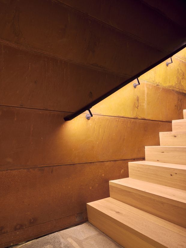 A close view of a warm, sculptural stairway with broad pale timber treads and risers ascending beside dark rust-brown metal or patinated feature walls. Slim black wall-mounted lighting washes the stair enclosure with amber light, creating a dramatic and tactile interior.