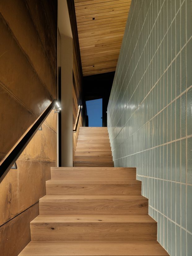 A narrow contemporary stairway rises through a tall, tightly enclosed passage. The stairs and ceiling are clad in warm natural timber, with a dark timber wall and integrated black handrail on the left, while the right wall is covered in pale blue-green glossy rectangular tiles with light grout. Small wall lights illuminate the steps, and a glimpse of blue sky is visible at the upper landing.