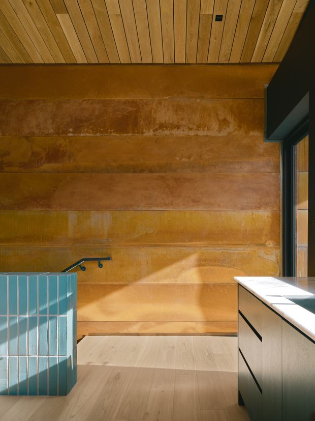 A close-up interior view of a contemporary kitchen, featuring warm rust-toned metal wall panelling, a light timber slatted ceiling, pale timber flooring, and a dark-framed window. The foreground includes a turquoise vertical tile splashback or partition and streamlined cabinetry with a light benchtop. Strong sunlight creates a warm, tactile, modern atmosphere.
