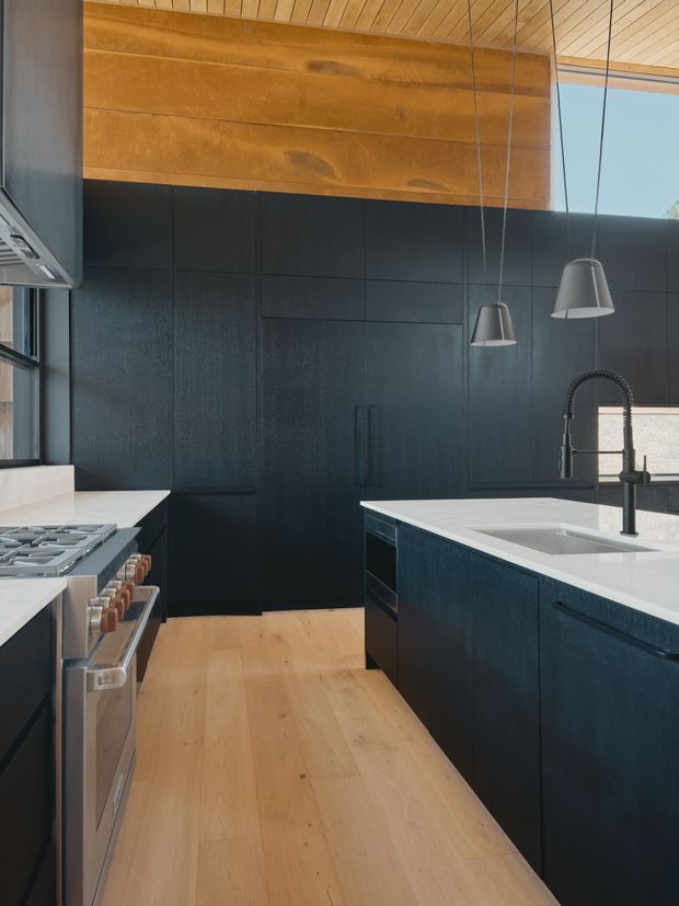 A contemporary kitchen with dark, vertically grained cabinetry, a large pale stone island benchtop and integrated sink, stainless-steel range and oven, and matte black spring-coil tapware. Warm timber wall panelling and a timber-lined ceiling contrast with the dark joinery, while pendant lights hang above the island. Timber flooring and large windows add natural light.