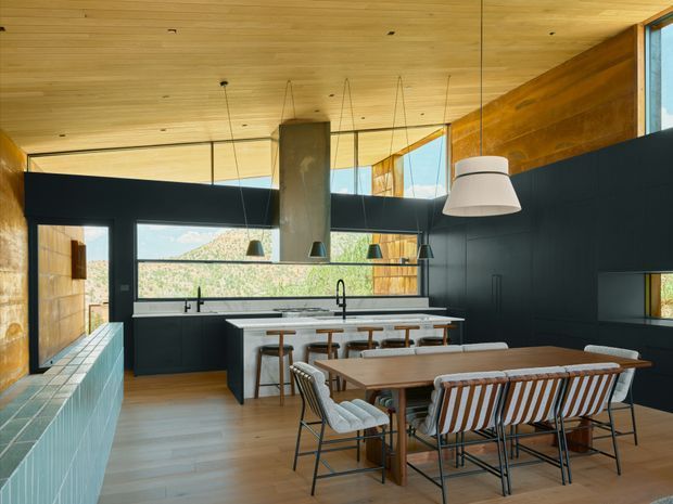 A spacious open-plan kitchen and dining area with dark matte cabinetry, a large white island, integrated cooktop and sink, and a central cylindrical rangehood. A timber dining table with upholstered chairs and a built-in bench occupies the foreground. Warm timber-lined ceilings and walls contrast with expansive black-framed glazing overlooking greenery, creating a contemporary, refined interior.