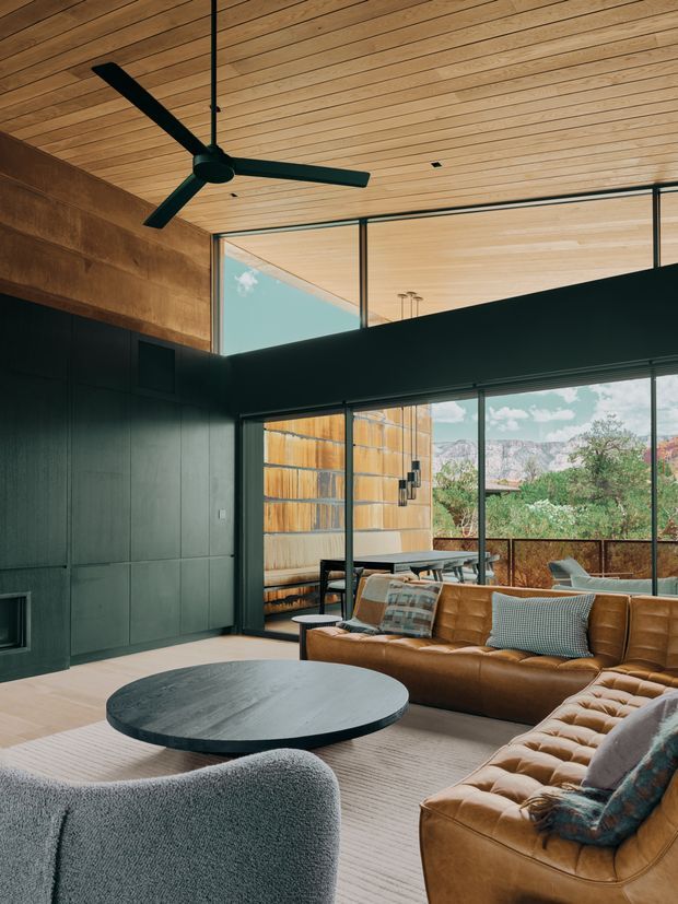 A warm, expansive living room with a high timber-lined ceiling, large black-framed windows, and a dark built-in wall. A cognac leather sectional surrounds a low round dark coffee table, with a textured lounge chair in the foreground. The glazing opens views toward a timber privacy fence, trees, and rocky hills, while a large black ceiling fan and subtle recessed lighting add a contemporary architectural character.