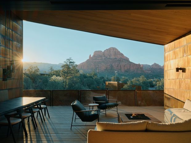A warmly lit contemporary outdoor living terrace framed by a deep timber-clad roof and walls, with a dining table, lounge seating, and a built-in fire feature. The open terrace overlooks dense vegetation and a dramatic red-rock mountain landscape, creating a strong indoor–outdoor connection.