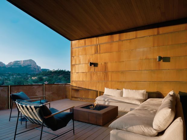 A contemporary outdoor terrace with timber decking, built-in upholstered seating, lounge chairs, and a square fire pit. Warm timber wall and ceiling cladding frame an open mountain-facing outlook, with a glass or metal balustrade along the edge. The space has a refined, resort-like outdoor living atmosphere.