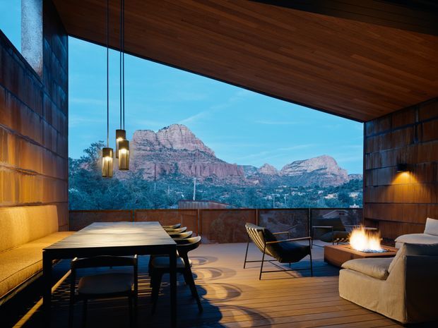 A sophisticated covered outdoor dining and lounge terrace framed by dark timber-clad walls and a broad timber soffit. The space includes a long dining table with upholstered bench seating, sculptural pendant lights, lounge chairs, a large sofa, and a fire feature. Full-height open glazing or an open-sided frame provides a dramatic view toward rugged red-rock mountains. The overall design is warm, refined, and strongly connected to the landscape.