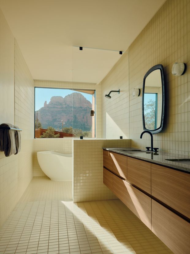 Bright contemporary bathroom with a freestanding white bathtub positioned beside a large window overlooking a rocky mountain landscape. The room features small cream square tiles across the walls and floor, a floating warm timber vanity with dark stone benchtop, black tapware and shower fittings, and a distinctive rounded rectangular black-framed mirror. Clear glass shower screening separates the bath area from the vanity. Strong natural sunlight creates a warm, refined atmosphere.