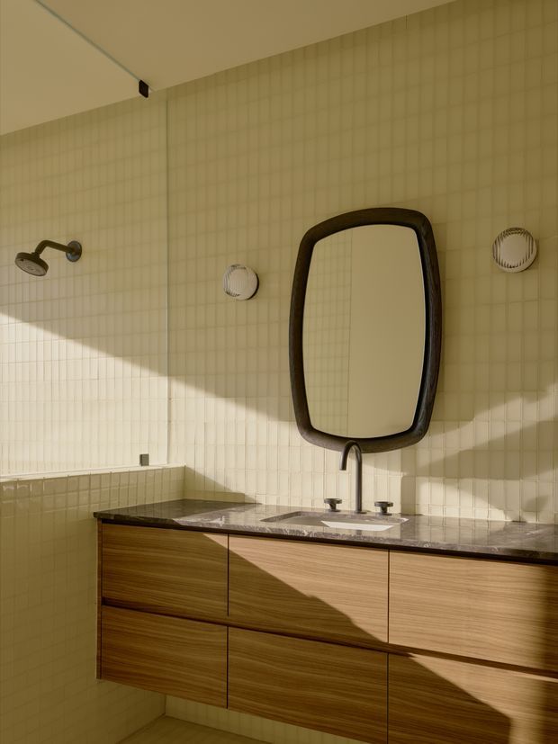 A warm, sunlit bathroom vanity with a long floating timber drawer unit, dark stone benchtop, integrated white basin, and brushed metal tapware. The wall is clad in small cream rectangular tiles, complemented by a large organic rounded-rectangle dark-framed mirror and a wall-mounted shower fitting. Strong diagonal sunlight creates dramatic shadows across the tiled wall and cabinetry.