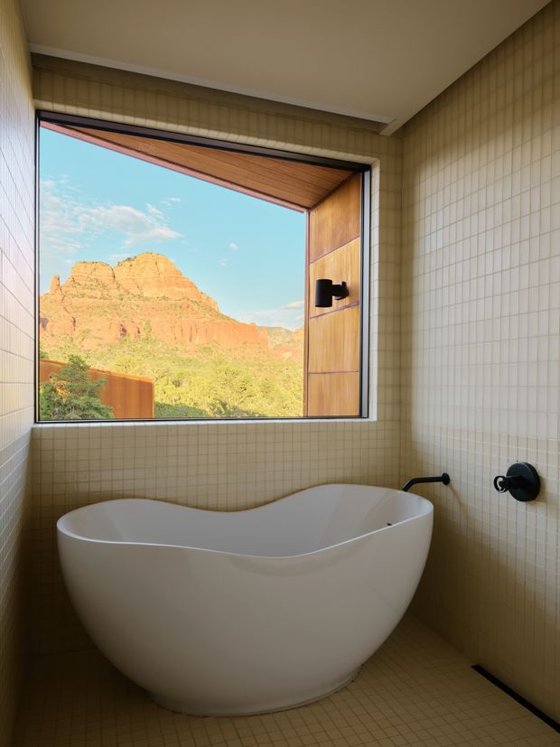 A bright contemporary bathroom featuring a freestanding white sculptural bathtub beneath a large open window. The walls and floor are covered in small beige square tiles, with matte black plumbing fixtures and a warm timber-lined window reveal. The window frames a dramatic red-rock desert landscape.
