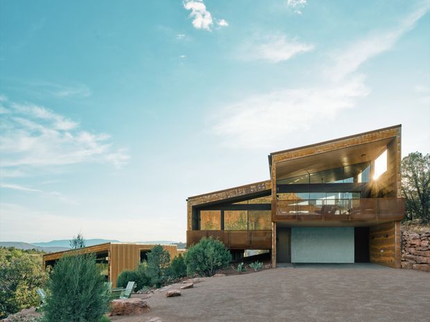 Exterior view of a contemporary multi-level residence on a sloping site, featuring projecting rectangular volumes, extensive glazing, warm timber-toned cladding and an integrated garage. A broad paved driveway and landscaped planting occupy the foreground, with distant hills visible beyond.