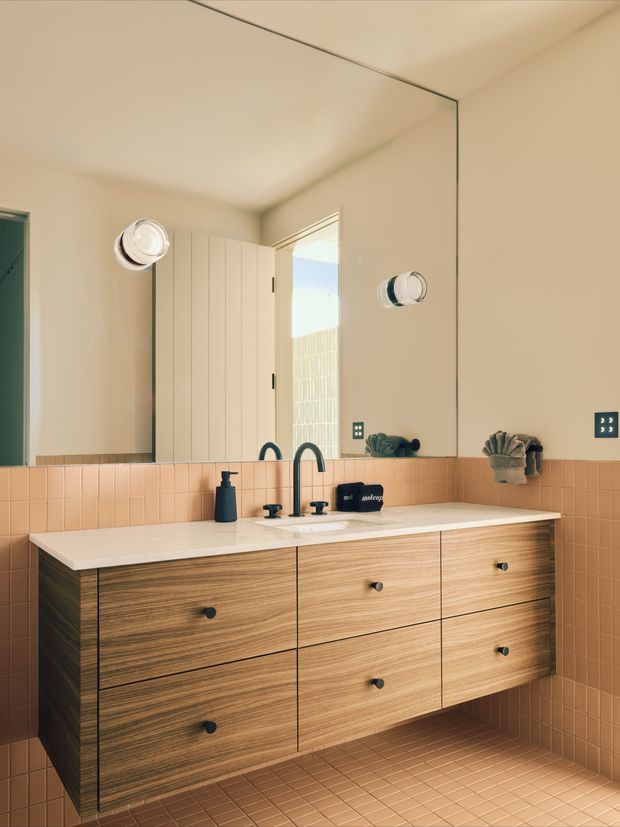 A warm contemporary bathroom vanity photographed straight on. The room features a large frameless mirror, a wall-mounted timber-grain floating vanity with multiple drawers, a pale stone or solid-surface benchtop, twin dark faucets and basins, and peach-pink square tiled walls and floor. An open door and small wall lights are reflected in the mirror.