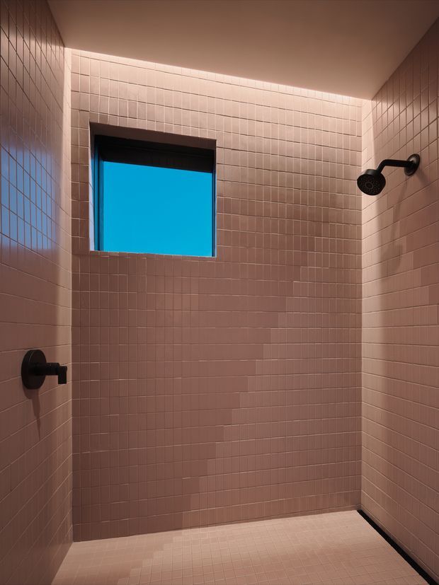 A compact, contemporary tiled shower room with blush-pink square tiles covering the walls and floor. The space features a black ceiling-mounted shower fixture, black control, dark window frame, recessed linear ceiling lighting, and a small rectangular window showing a bright blue exterior view. The image is architectural and largely unfurnished, emphasizing the tilework and fixtures.