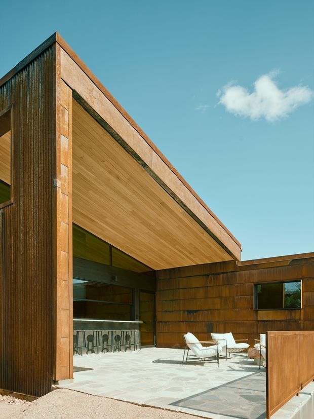 Exterior architectural view of a contemporary, angular building with a large cantilevered roof and open-air terrace. The facade combines vertically ribbed dark timber cladding with warm weathered wood panels, while the soffit is lined with lighter timber. A paved outdoor seating area, bar counter, and glazed openings sit beneath the deep roof overhang. The image is brightly lit under a clear blue sky.