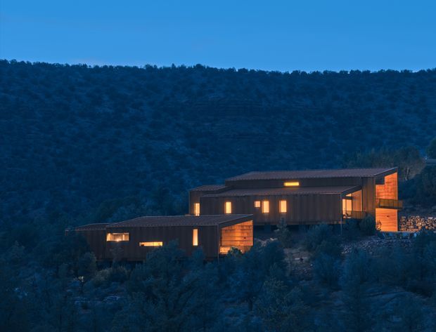 A low, contemporary multi-volume house set into a wooded hillside at dusk. The dark horizontal building forms have shallow pitched or nearly flat roofs, extensive warm-lit glazing, and timber-toned wall sections. Dense bush surrounds the residence, with a rugged stone retaining wall visible at the right.