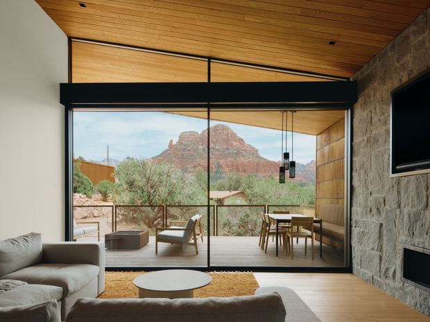 A warm, modern living room with a large floor-to-ceiling glazed opening overlooking a dramatic red-rock landscape. The room features a timber-lined ceiling and wall, pale timber flooring, a substantial rough stone feature wall with built-in fireplace and television, neutral upholstered sofas, a low round coffee table, and a dining area opening onto a timber deck. Black metal-framed glazing creates a strong horizontal architectural emphasis.