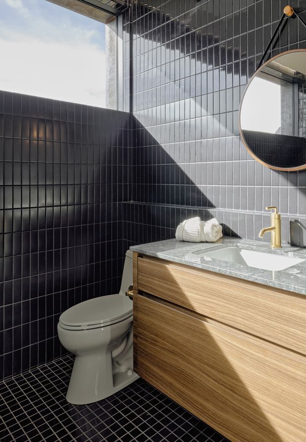 Compact contemporary bathroom with black tiled walls and floor, a timber vanity with a pale stone benchtop, white basin and toilet, brass tapware, and a round framed mirror. Strong natural light enters from above or through glazing, creating geometric shadows across the dark tiled surfaces and vanity.