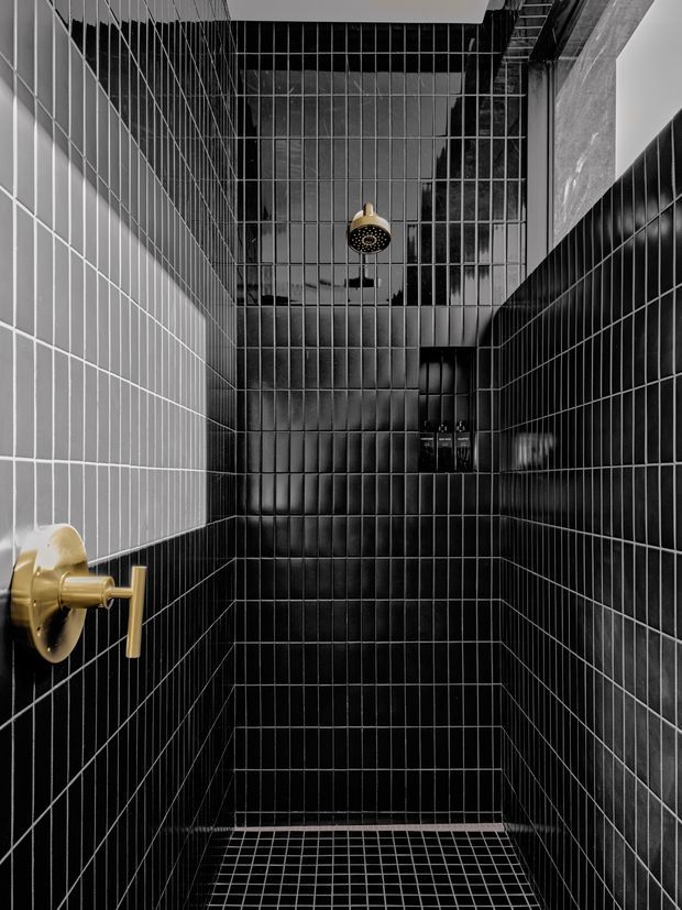 A compact, dramatic walk-in shower lined floor-to-ceiling with glossy black rectangular tiles and a matching small-format black tiled floor. The shower features a gold/brass rain showerhead and matching control hardware, with a recessed niche holding toiletries. A bright vertical opening or window is visible near the upper right, contrasting with the dark tiled enclosure.