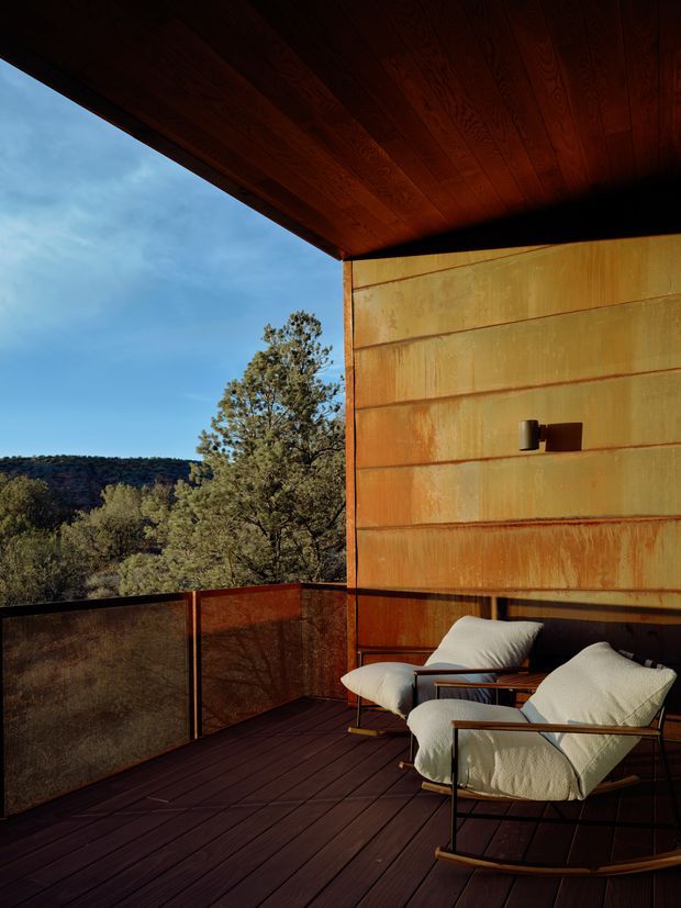 A covered outdoor terrace overlooking a wooded, hilly landscape. The space features dark timber decking and a timber-lined soffit, a rust-toned weathered metal wall and balustrade, and two cream upholstered lounge chairs with dark metal frames. Warm late-day light creates a dramatic, secluded retreat-like atmosphere.