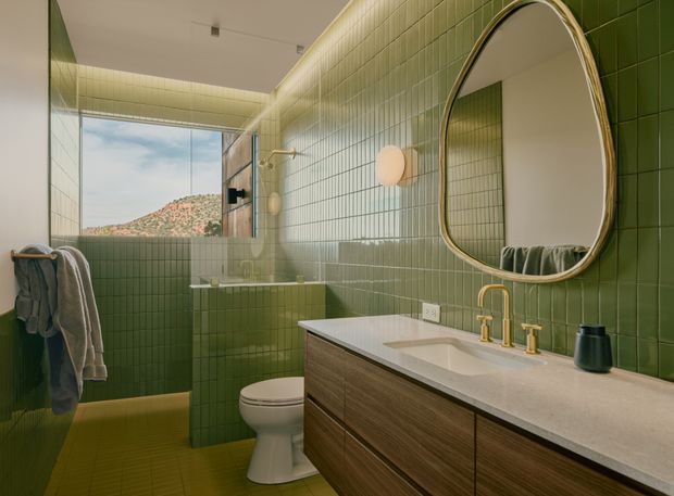 A warm, contemporary bathroom clad in small glossy green tiles, featuring a wood-toned floating vanity with a pale stone benchtop, integrated basin, brushed gold tapware, and a large rounded-oval mirror. A tiled shower and freestanding toilet sit beside a floor-to-ceiling window overlooking a rocky hillside. A towel rail and soft wall lighting complete the space.