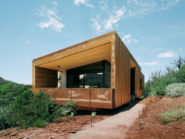 A compact contemporary pavilion-like dwelling set within a natural, scrub-covered landscape. The rectilinear structure features weathered rust-toned metal cladding, warm timber soffits and wall surfaces, extensive dark-framed glazing, and a recessed covered outdoor terrace with a transparent balustrade. A narrow path approaches the elevated building through reddish rocky terrain under a bright blue sky.