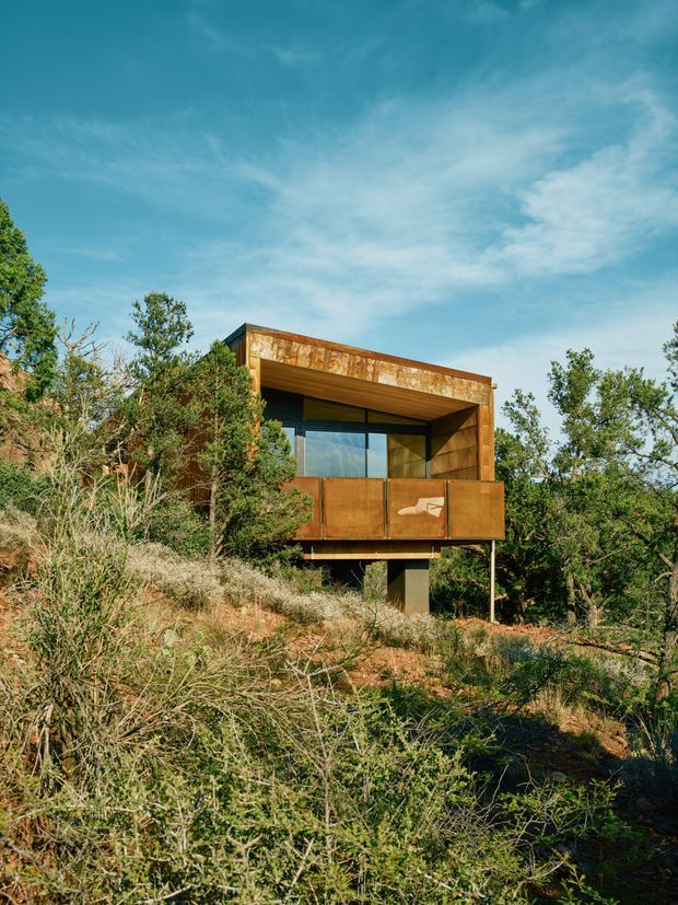 Exterior view of a compact elevated dwelling set within a dry, scrubby natural landscape. The building has a strongly rectilinear form, deep covered balcony or terrace, large glazing, and weathered rust-toned metal cladding. It is supported on narrow posts above the sloping site and is partially screened by native vegetation.