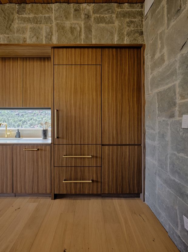A warm, compact kitchen interior featuring full-height dark timber cabinetry with integrated drawers and brass-toned handles, a light stone benchtop, and a small window above the sink. Exposed rough-cut stone masonry forms the surrounding walls, while wide timber floorboards and a timber-lined ceiling reinforce the natural, earthy character. A green, wooded landscape is visible through the window.