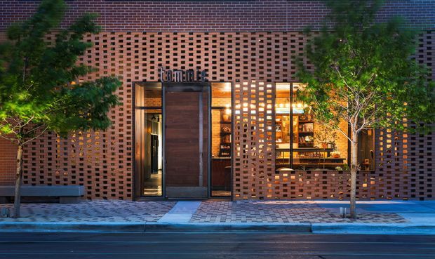 Street-facing commercial storefront at dusk, composed of warm red-brown brickwork with extensive perforated brick screens around a dark timber or wood-toned entry door and illuminated display windows. Small street trees, patterned paving, and warm interior lighting create a refined urban facade.