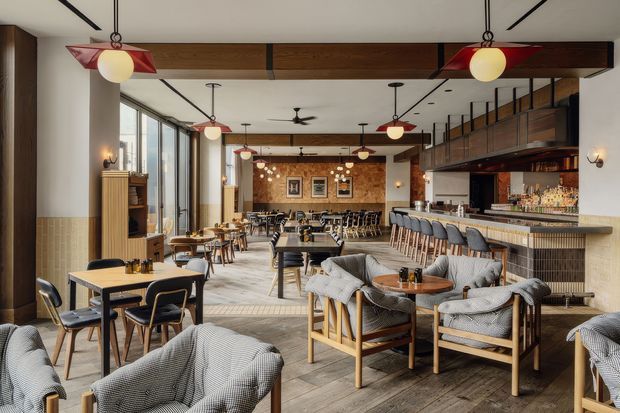 A spacious, warmly styled restaurant or hospitality venue with a bar along the right side, extensive dining seating, lounge chairs in the foreground, and a long open dining area extending toward a textured feature wall. The interior combines timber beams and flooring, cream walls, tiled lower wall surfaces, upholstered seating, decorative pendant lighting, and large windows along the left side.