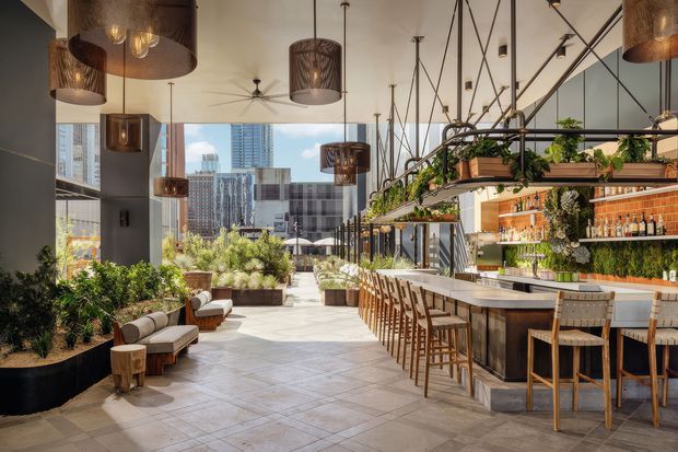 A bright, spacious contemporary rooftop bar or hospitality venue with a long stone-topped counter, timber bar stools, extensive hanging greenery, and illuminated shelving displaying bottles and glassware. The room has large floor-to-ceiling windows overlooking a dense urban skyline, suspended woven pendant lights, polished concrete columns, and a light tiled floor. Lounge seating and planted areas line the opposite side of the open space.